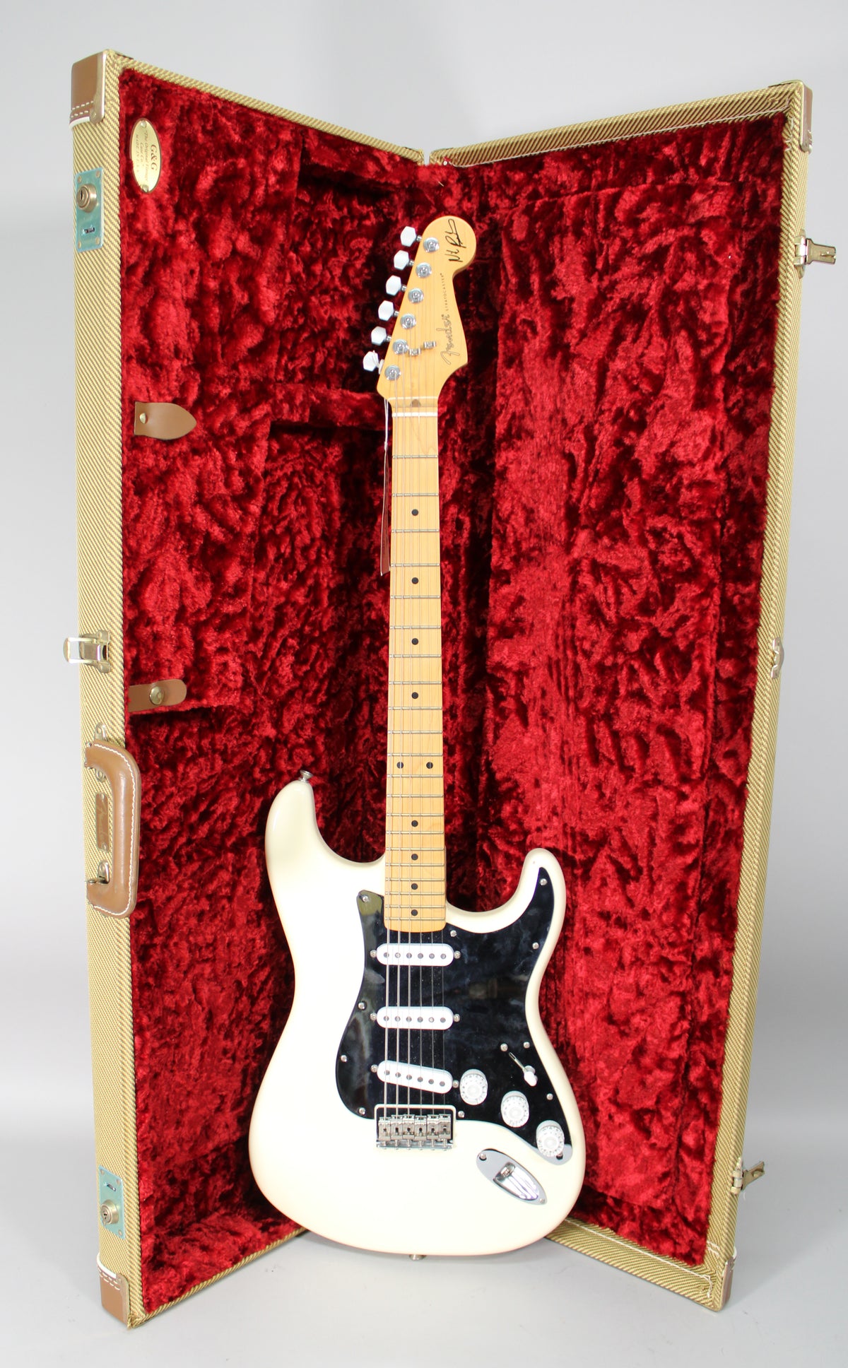 Olympic white Stratocaster with a black pickguard, three single-coil pickups, and a maple neck, standing in a tweed hard case with red velvet lining.