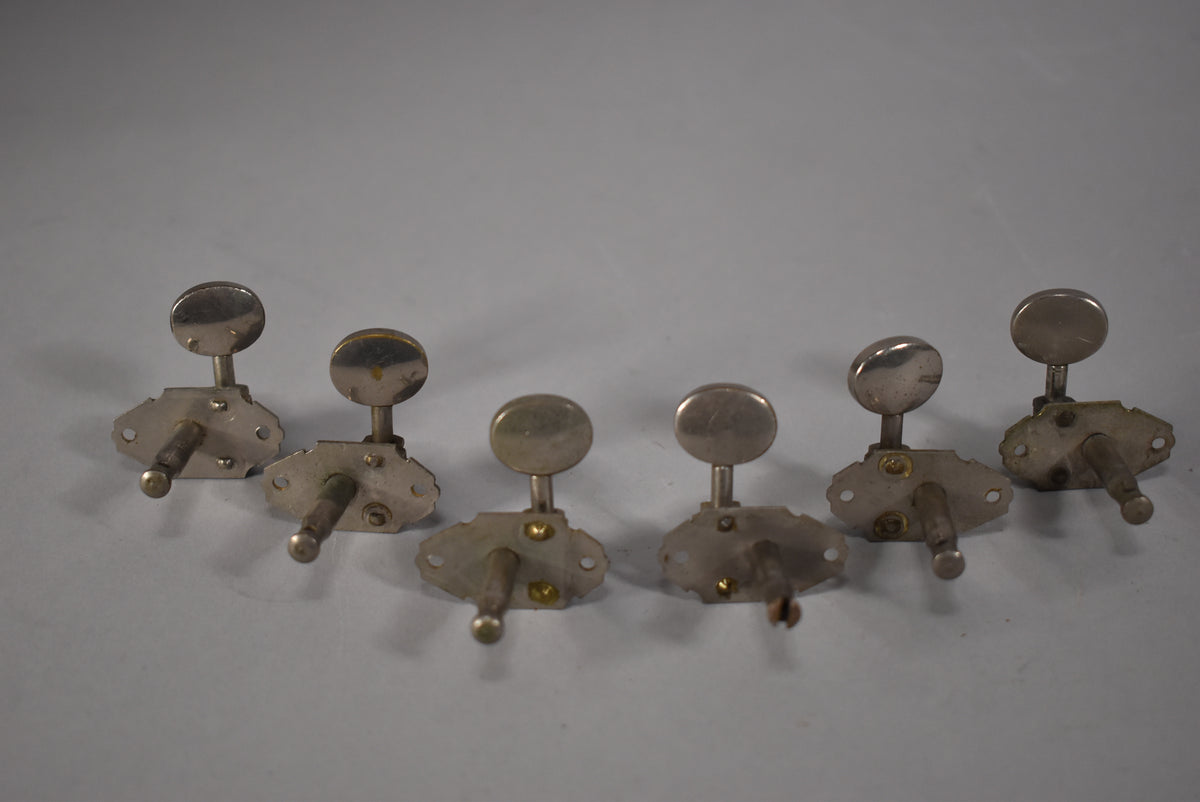 Six tarnished metal guitar tuners with oval buttons and exposed gears are arranged in two rows of three on a light gray surface.