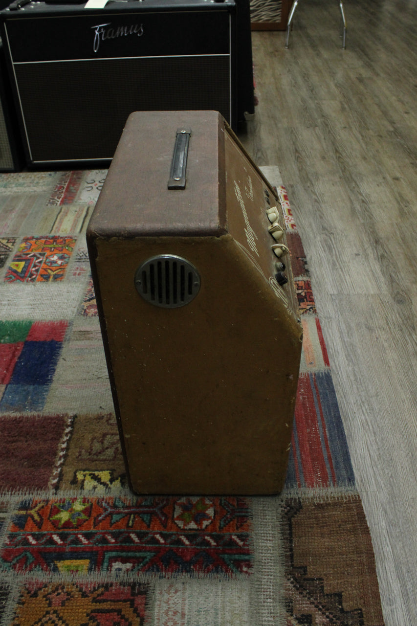 The brown tweed amp is angled from the top right, showing a metal handle, a round vent, and four cream control knobs.
