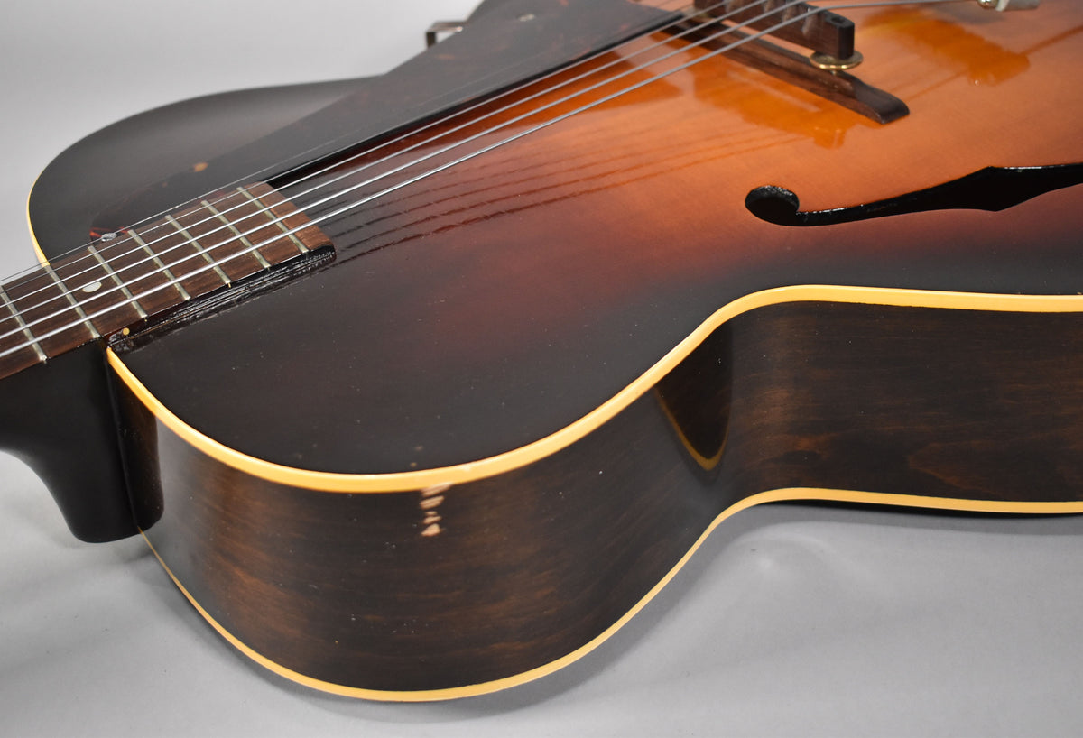 The sunburst archtop's lower bout and side are visible, showing a dark brown finish with light binding and some wear on the side.