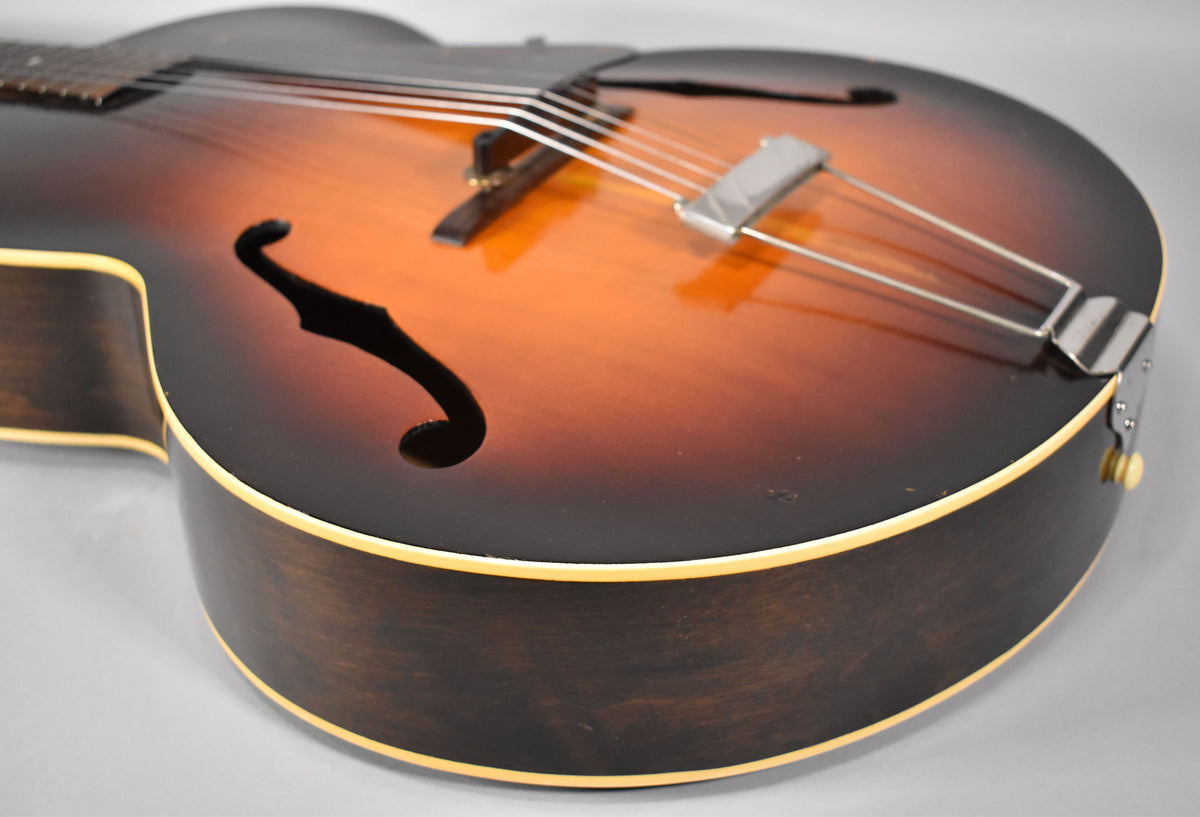 The sunburst archtop's lower bout and F-hole are visible, showing a chrome trapeze tailpiece and light wear on the dark sides.