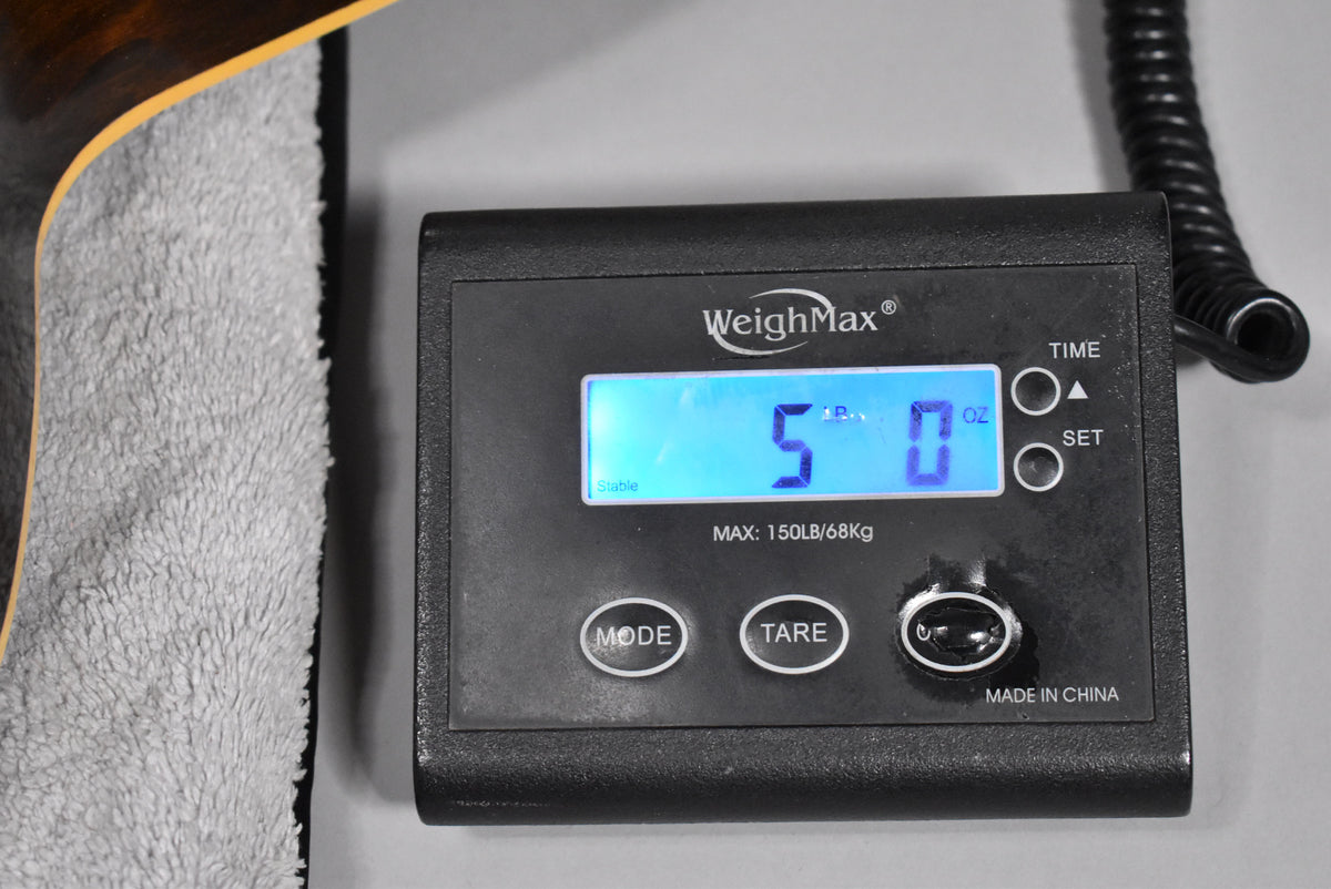 The guitar's dark sunburst body edge and binding rest on a grey towel, next to a digital scale displaying "5 lb 0 oz".