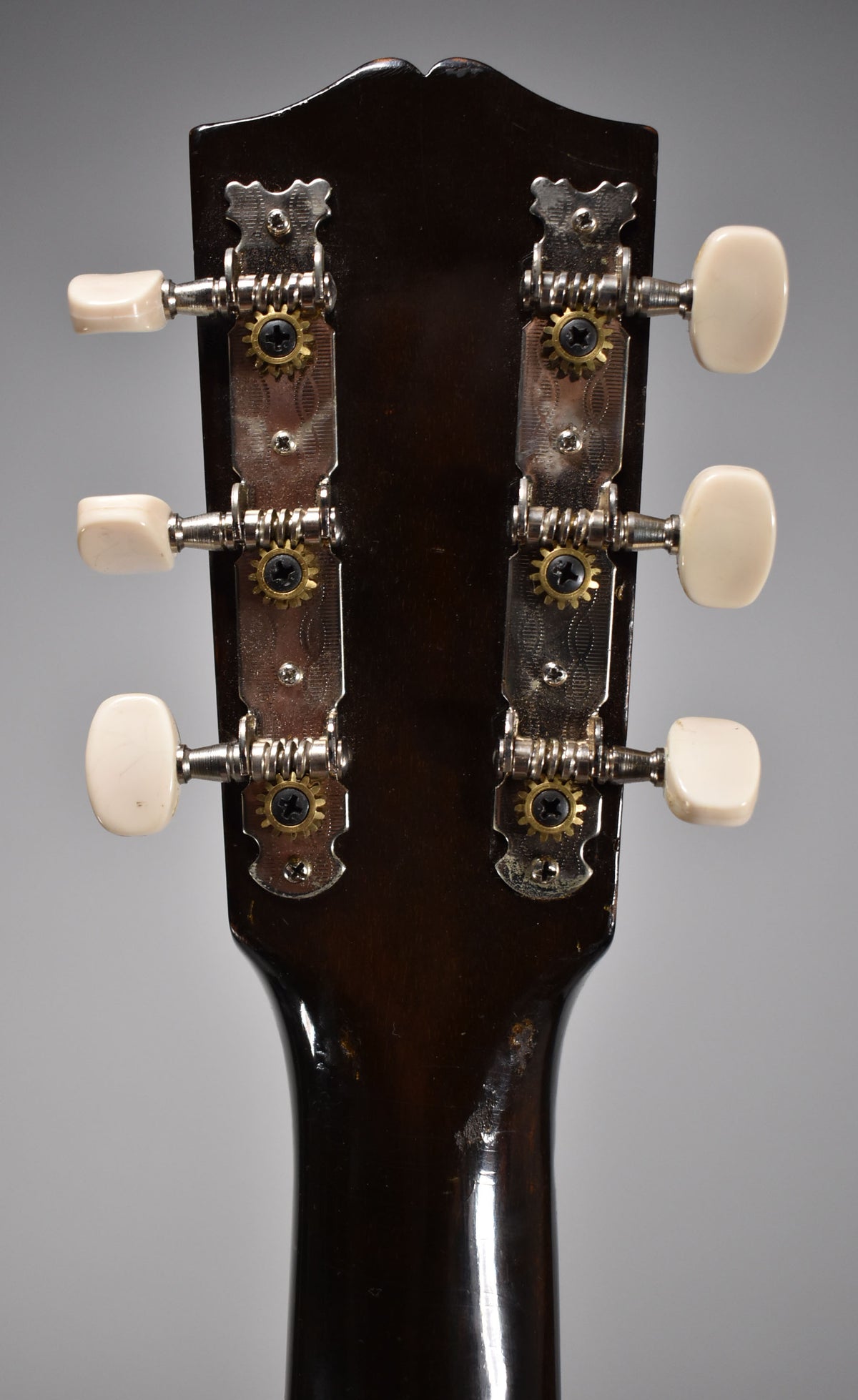 The dark brown headstock's rear shows six cream tuning pegs on two chrome plates with visible gears, and some finish wear on the neck.