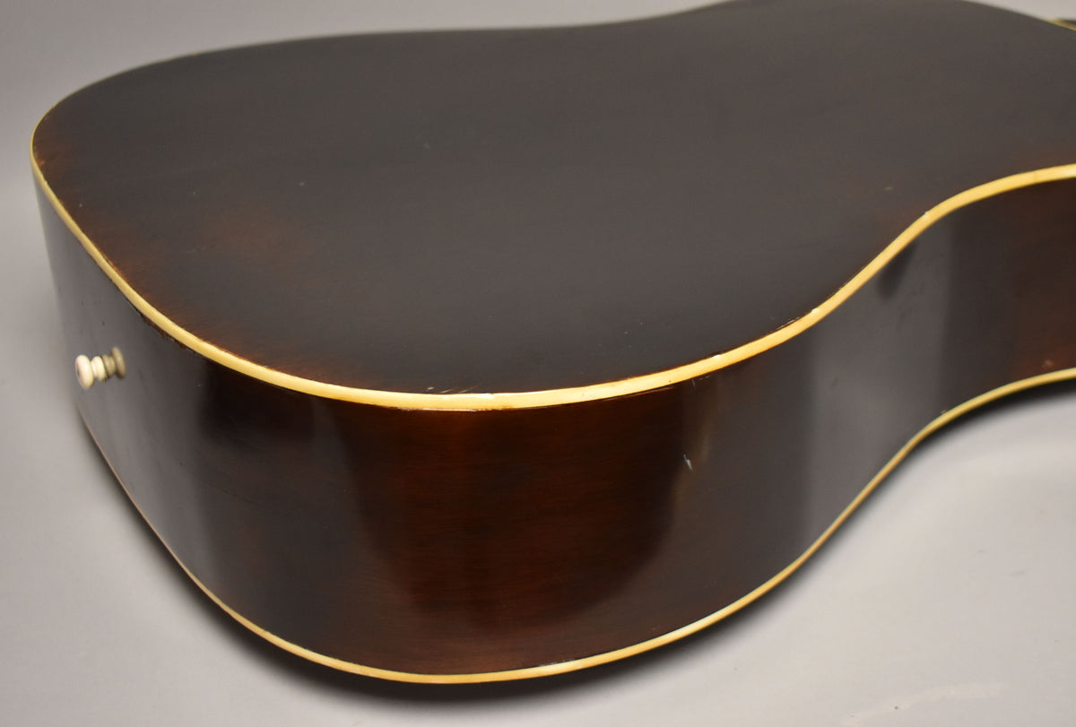 The lower bout of the dark sunburst acoustic guitar, showing the binding and a white end pin, with minor wear on the finish.