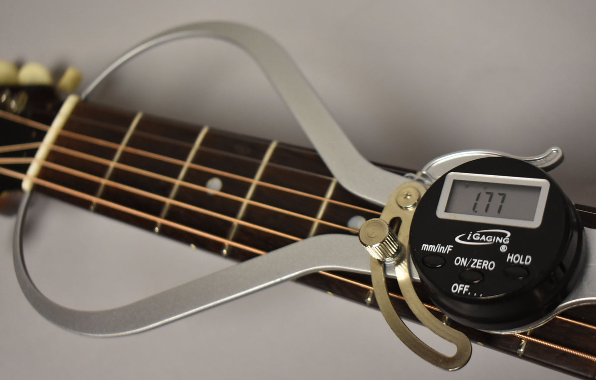 A digital caliper measures the guitar's dark fretboard, showing "1.77" on its screen, with light strings and dot inlays visible.