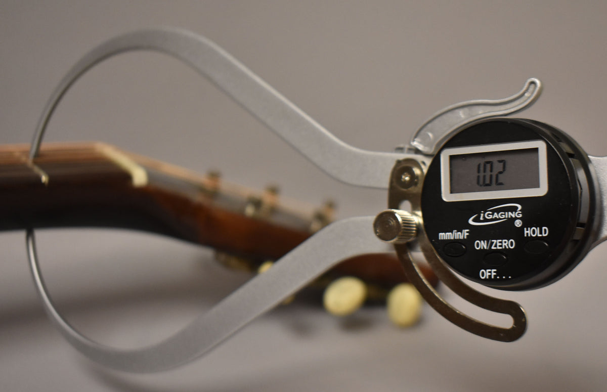 A digital caliper measures the guitar's neck thickness at the nut, displaying "1.02" with tuning pegs visible in the background.