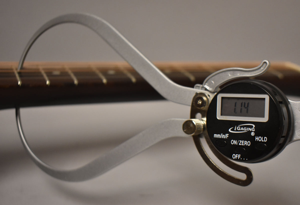 A digital caliper measures the guitar's dark brown fretboard, showing a reading of 1.14, with light wear on the frets.