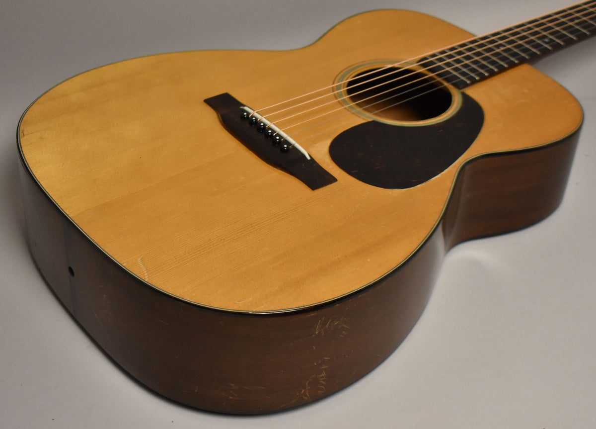 The natural finish acoustic guitar is angled from the lower bout, showing a black pickguard, bridge, and heavy wear on the dark sides.