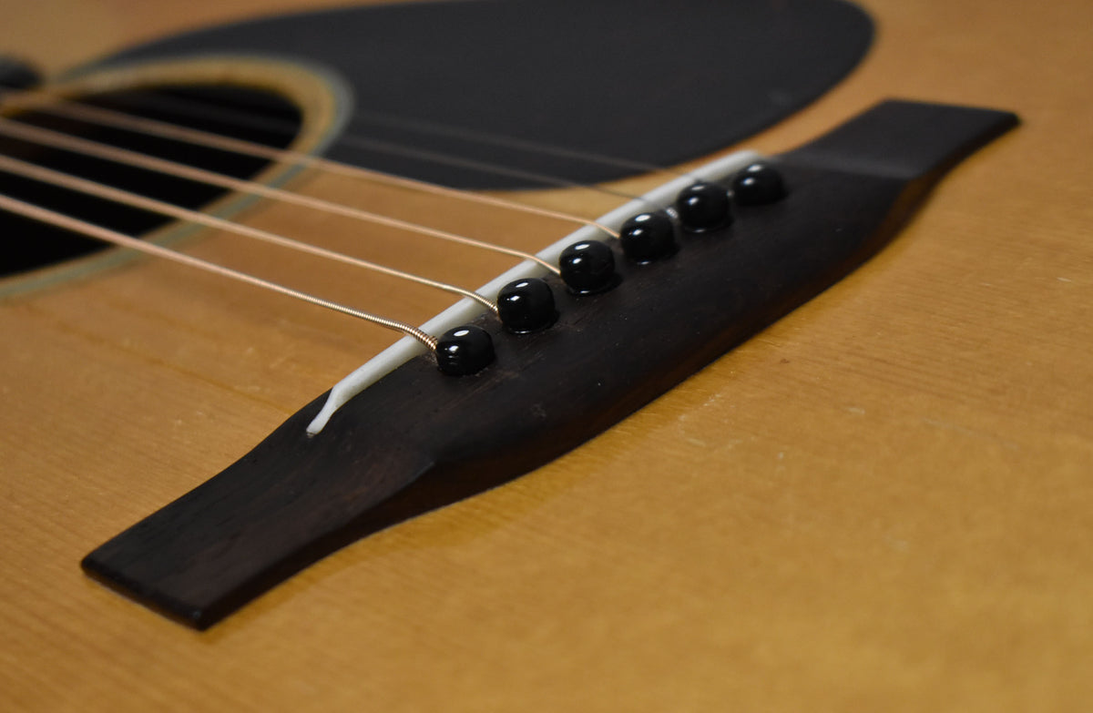 Close-up of the guitar's natural-finish body, showing the dark bridge, white saddle, six black bridge pins, and light-colored strings.