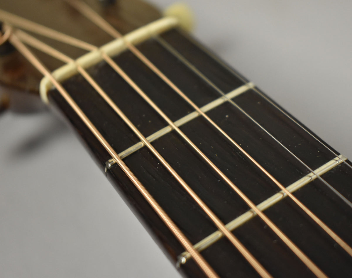 Close-up of the dark fretboard and light frets, with six bronze strings running over a light-colored nut.