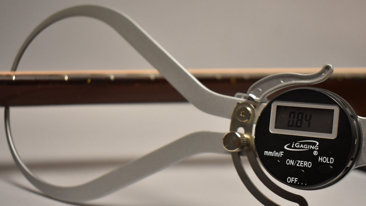 A digital caliper measures the dark brown neck of the guitar, showing 0.84 on its display.