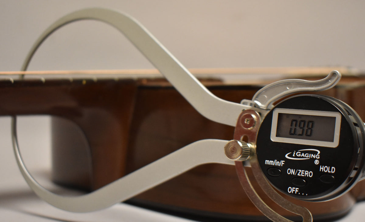 A digital caliper measures the string height above the fretboard, showing "0.98" on its display, against the guitar's dark brown body.