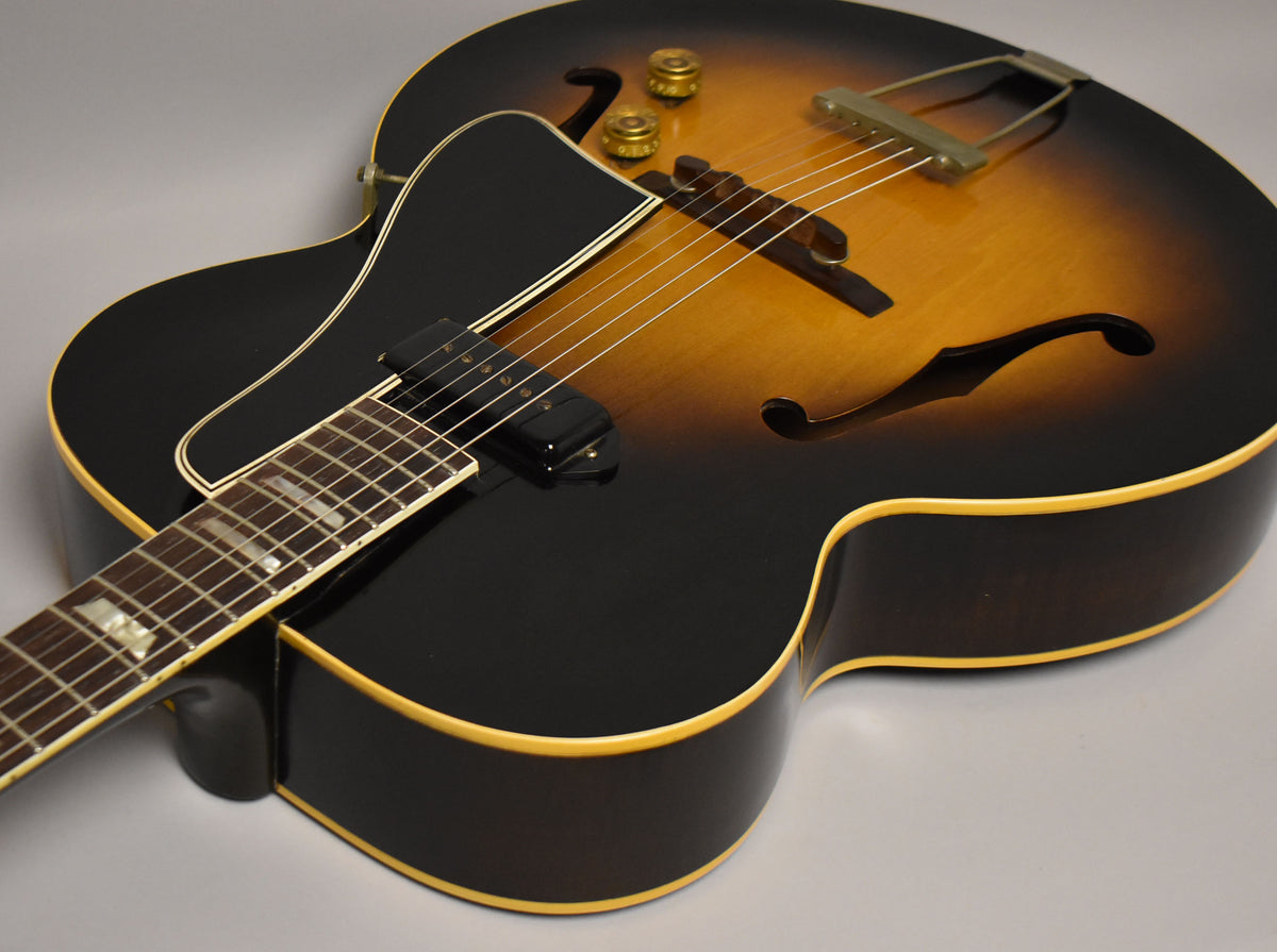 The sunburst hollowbody guitar is angled from the lower left, showing the dark brown sides, black pickguard, and f-holes.