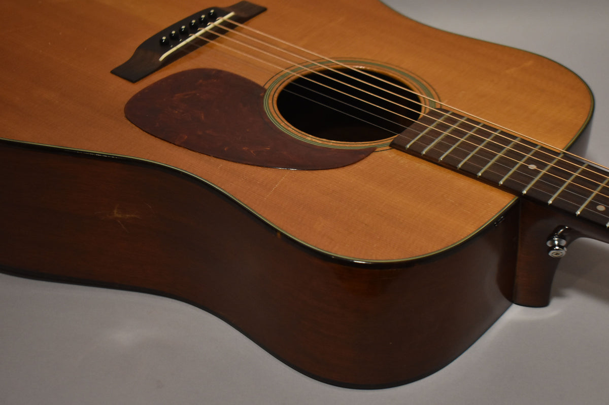 The natural finish acoustic guitar is angled to show the body, soundhole, pickguard, and lower fretboard, with visible wear and checking.
