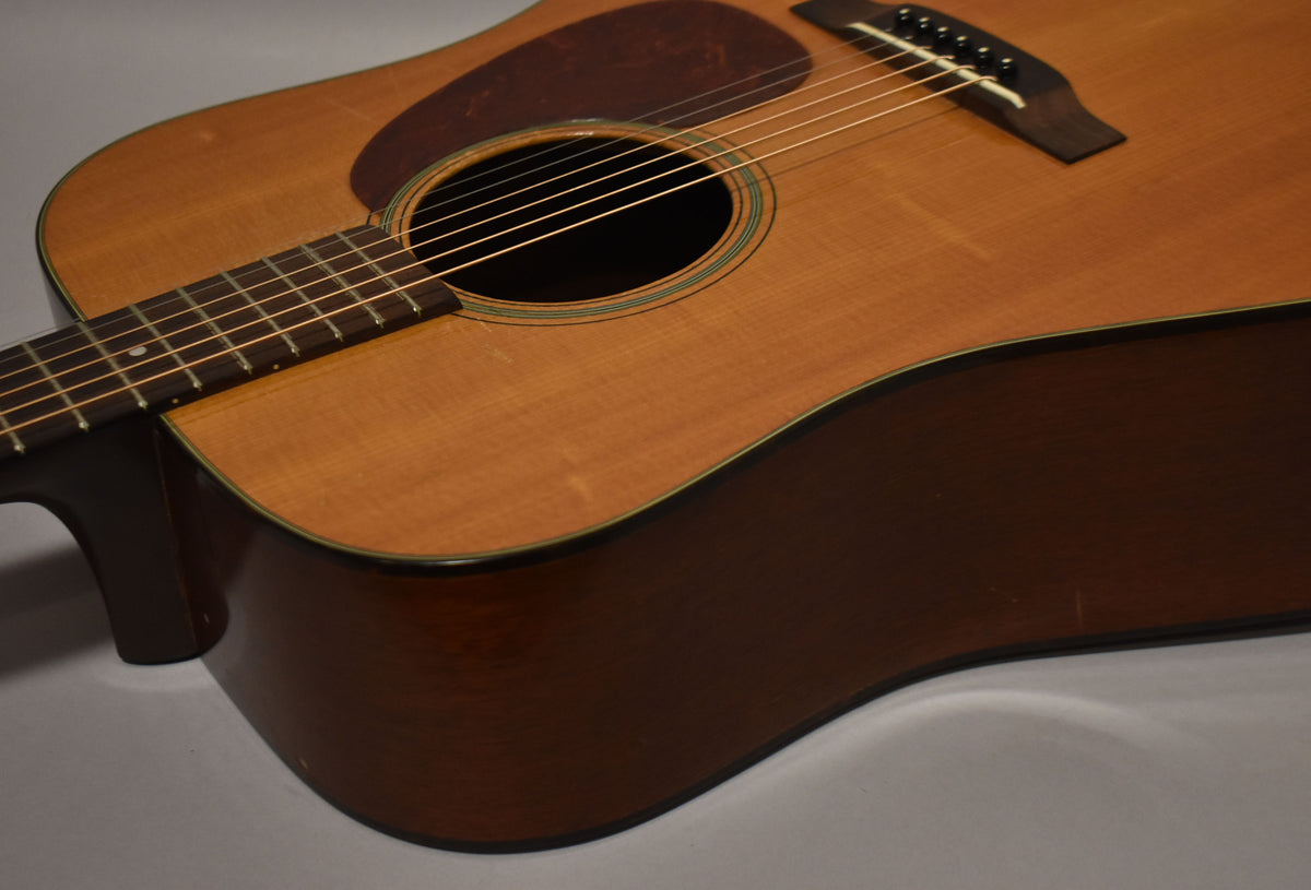 The natural finish acoustic guitar is angled, showing its light brown top, dark brown sides, pickguard, soundhole, and fretboard.