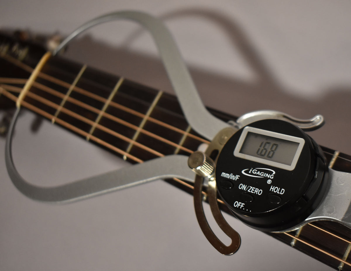 A digital caliper measures the guitar's nut width at 1.68 inches, resting on the dark fretboard with light strings.