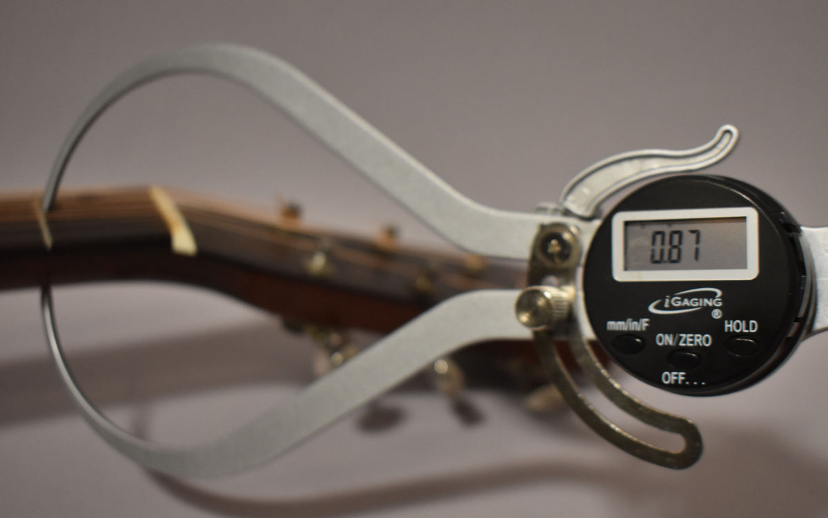A digital caliper measures the guitar's neck thickness at 0.87 inches, with the dark brown neck and light fretboard visible.