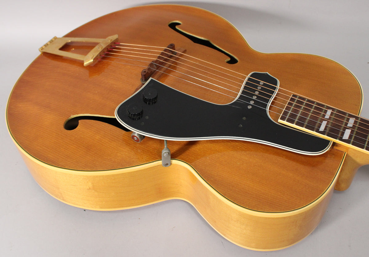 Upper body of a natural finish archtop, showing the black pickguard with two knobs, f-holes, and gold trapeze tailpiece.