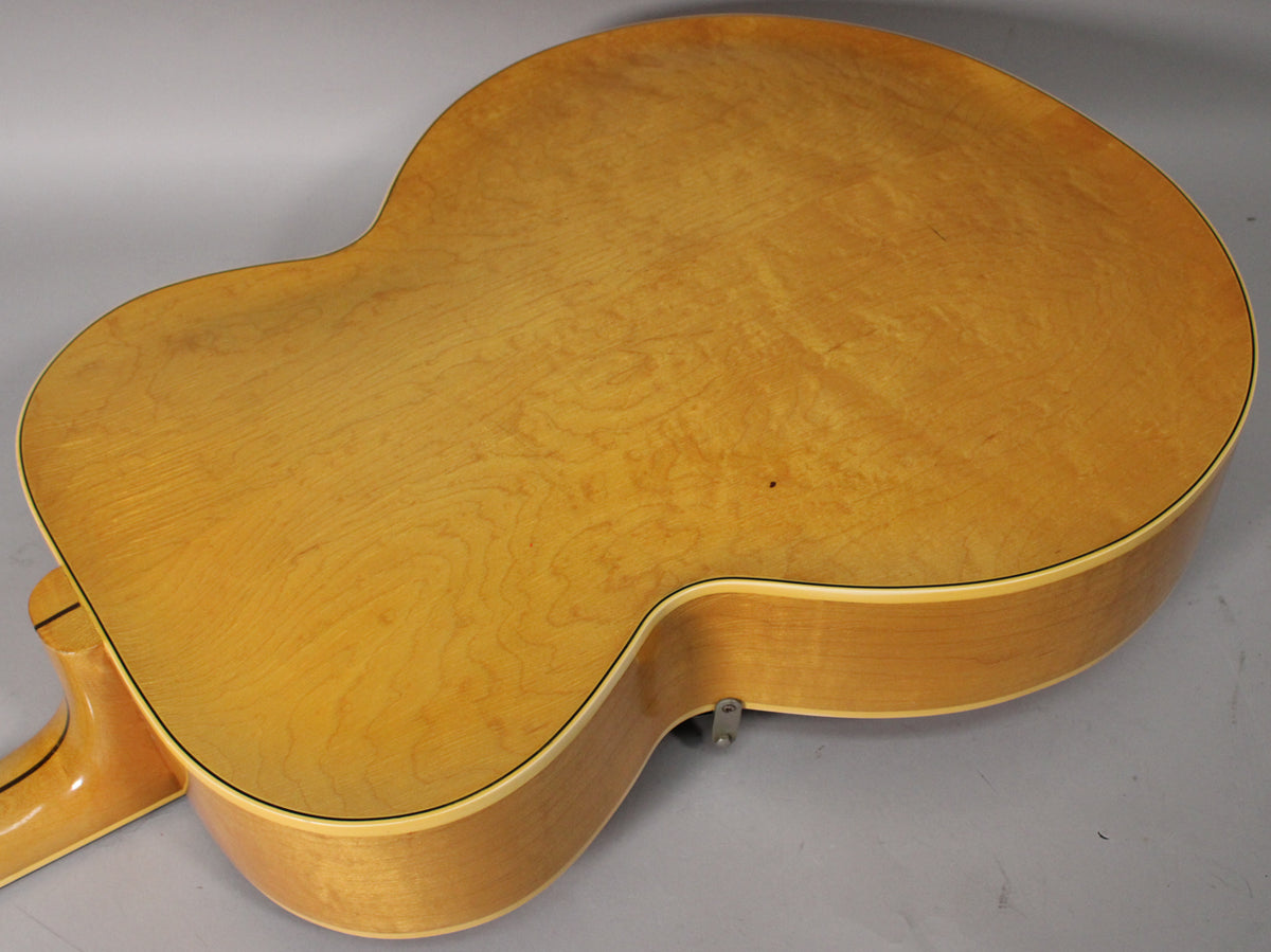 The back of the natural finish archtop guitar, showing light wood grain, a small dark spot, and a strap button on the lower bout.
