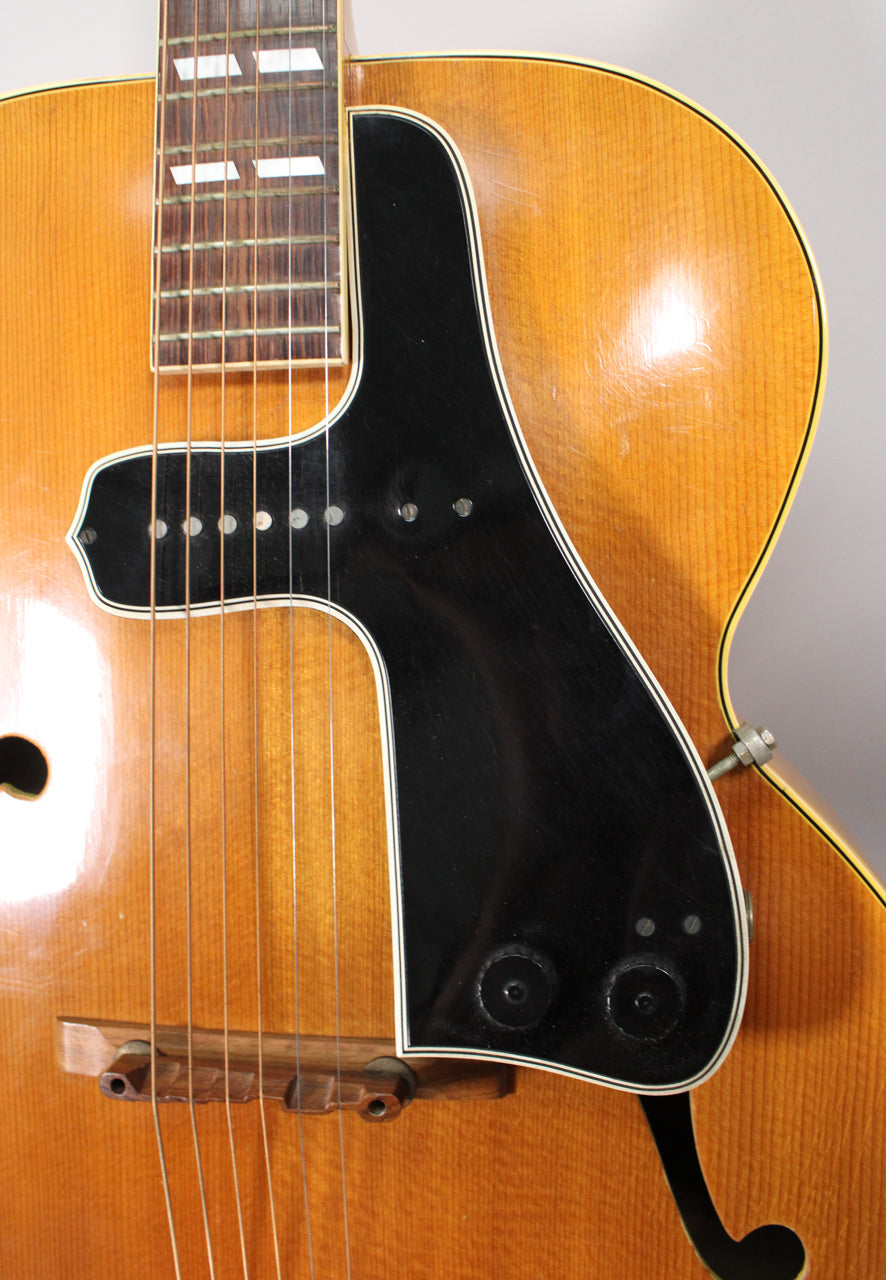 Close-up of the guitar's natural finish body, showing the black pickguard with two control knobs, a single pickup, and a wooden bridge.