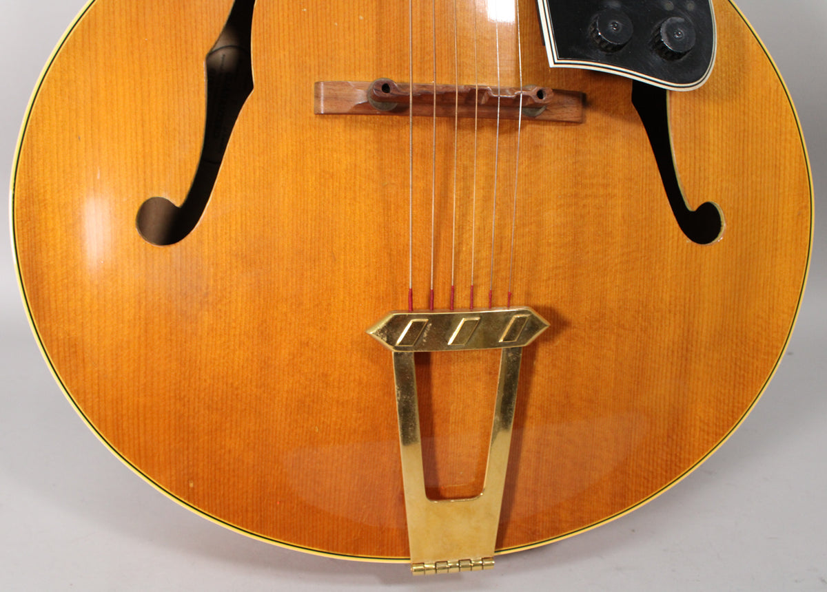 Close-up of the guitar's natural finish body, showing the f-holes, wooden bridge, gold trapeze tailpiece, and black pickguard with controls.