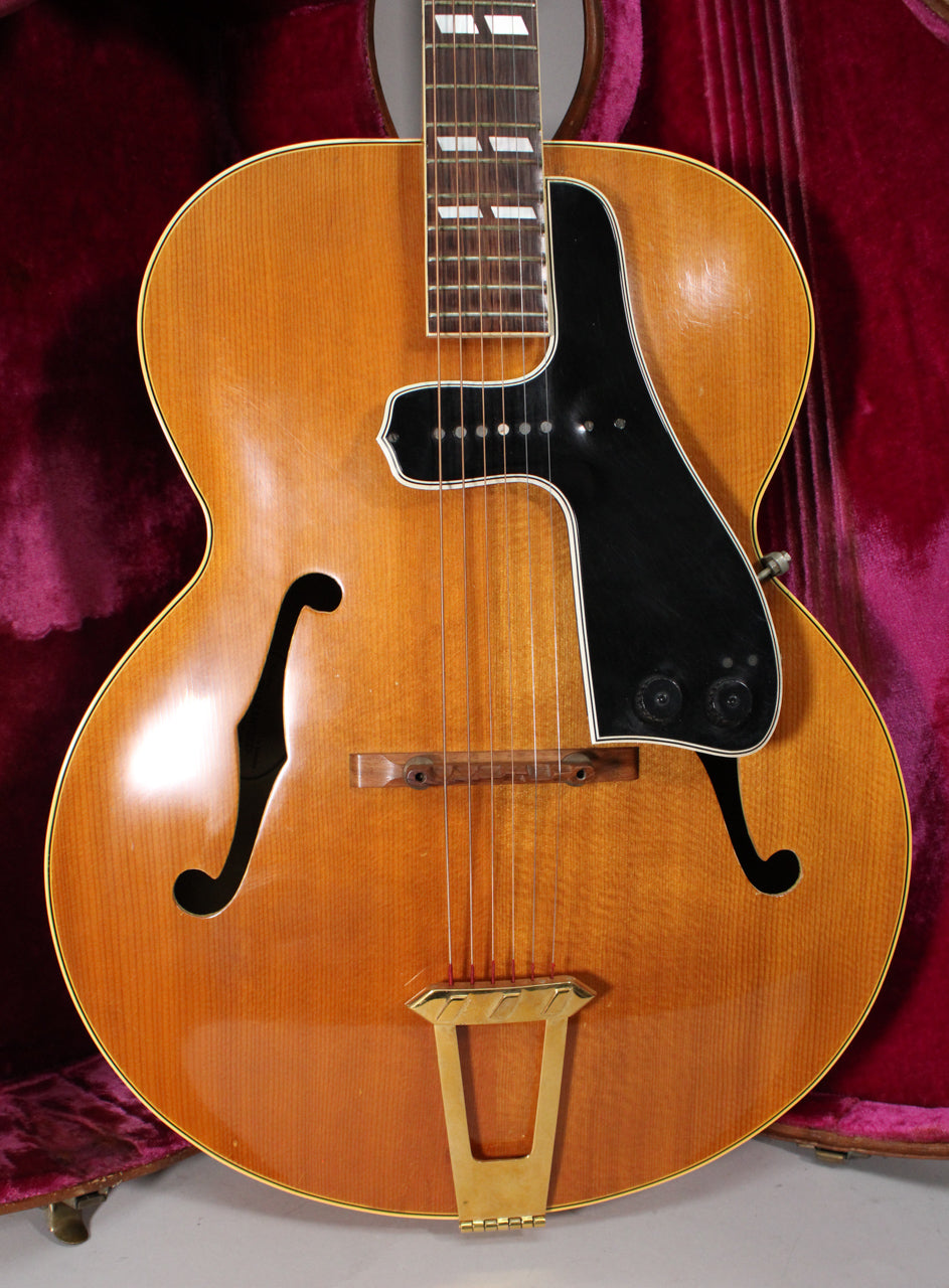 Full front view of a natural finish archtop guitar with f-holes, a black pickguard, two control knobs, and a gold trapeze tailpiece.