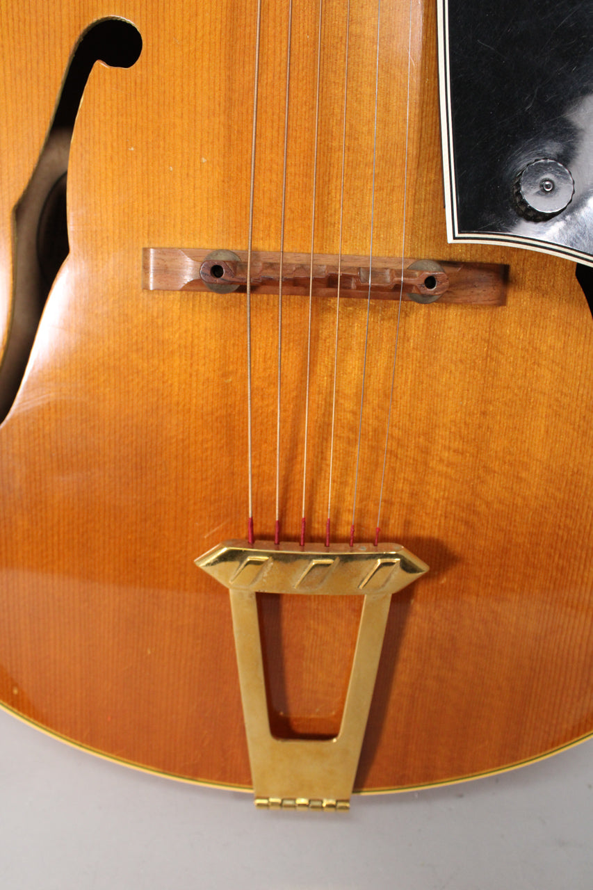 Close-up of the guitar's lower body, showing the natural finish, wooden bridge, gold tailpiece, and black pickguard with a control knob.