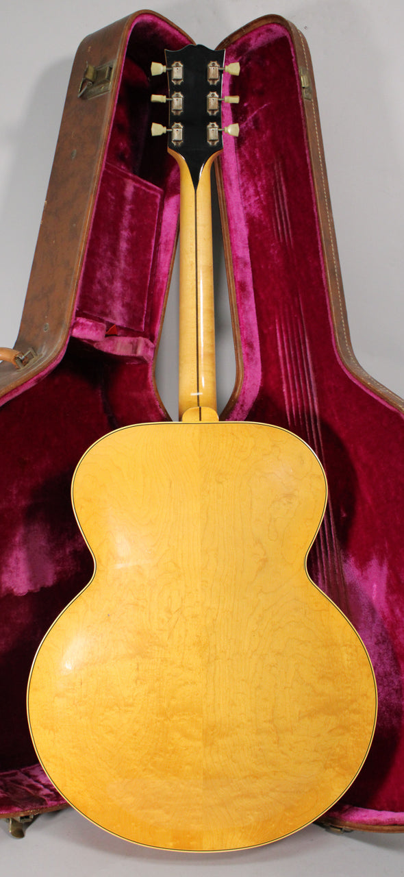 The back of a light blonde archtop guitar with a black headstock and cream tuners, resting in a red velvet-lined brown case.