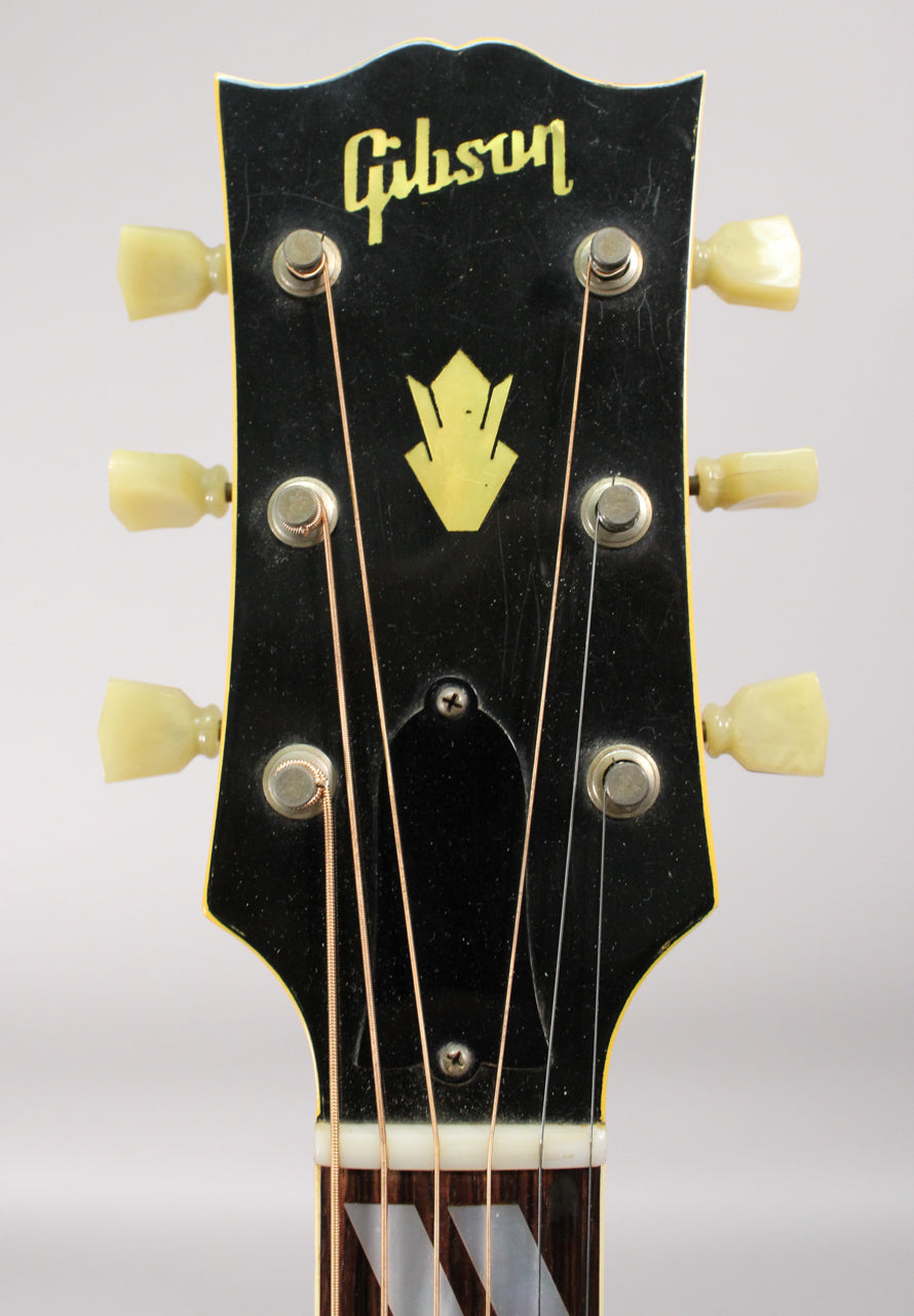 Close-up of the guitar's black headstock with a gold logo, six cream tuning pegs, and a partial view of the fretboard.
