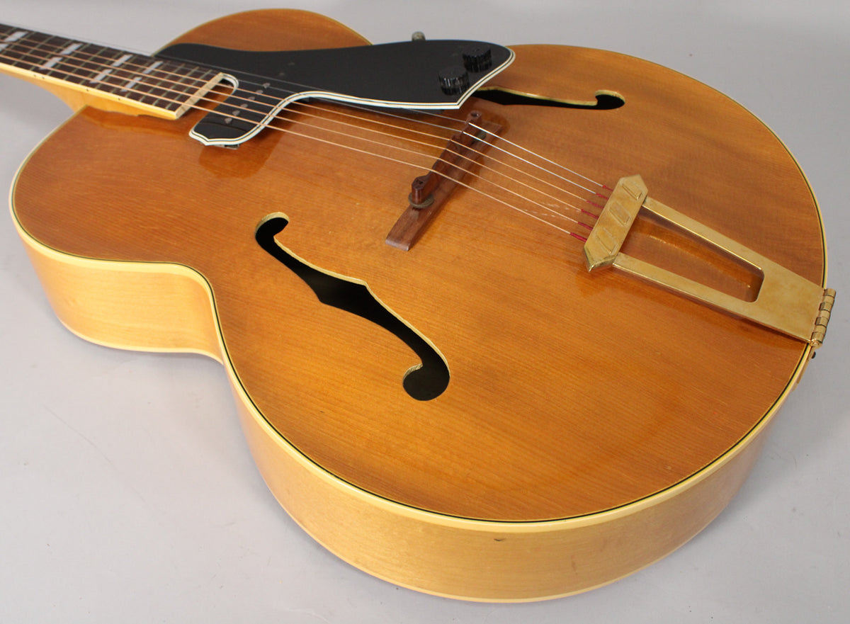 Close-up of the natural finish archtop guitar body, showing the black pickguard with two control knobs, f-holes, and a gold tailpiece.