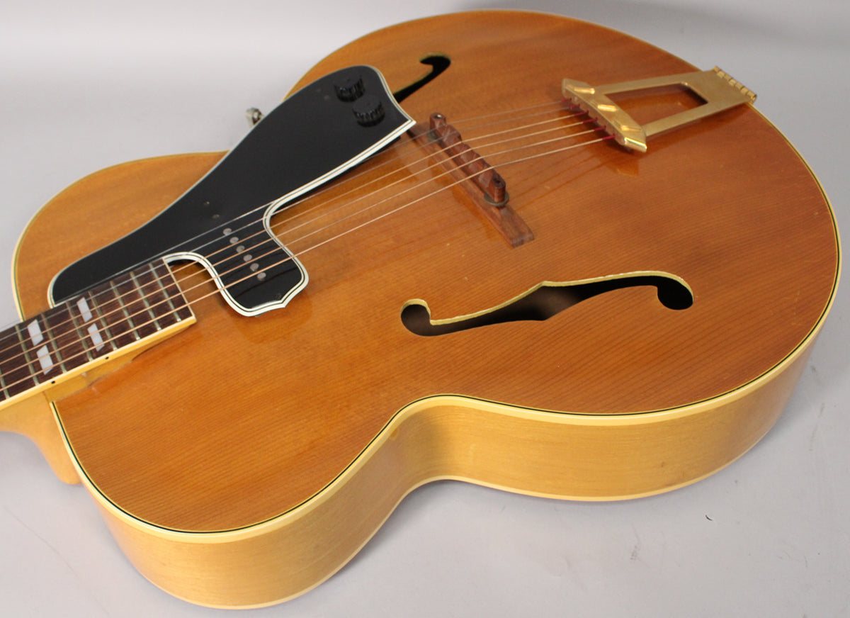 The natural finish archtop guitar is angled right, showing the upper bout, f-holes, black pickguard with controls, and gold tailpiece.