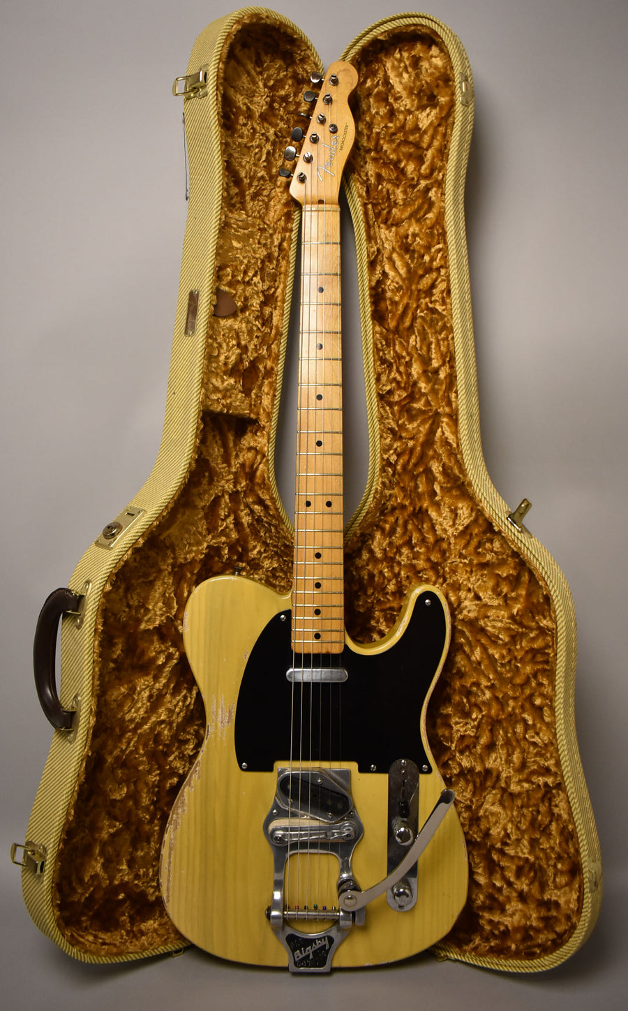 Full-length view of a butterscotch blonde electric guitar with a black pickguard, Bigsby tremolo, and heavy finish wear, standing in its tweed hard case.