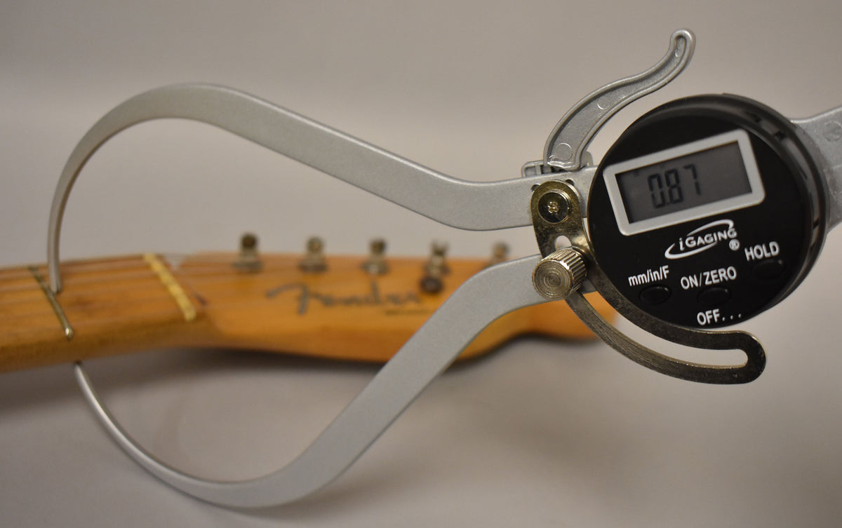 A digital caliper measures the guitar's neck thickness at the first fret, showing 0.87 inches, with the headstock blurred in the background.
