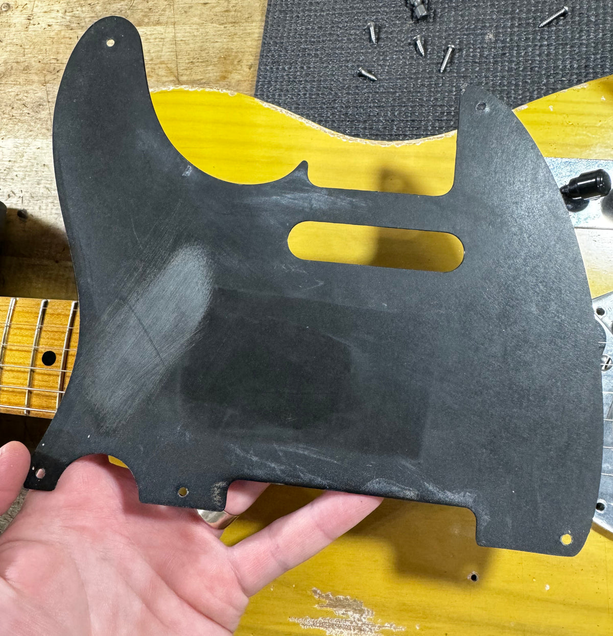 A hand holds the black pickguard, showing its worn, matte finish and two rectangular cutouts, above the guitar's blonde body.