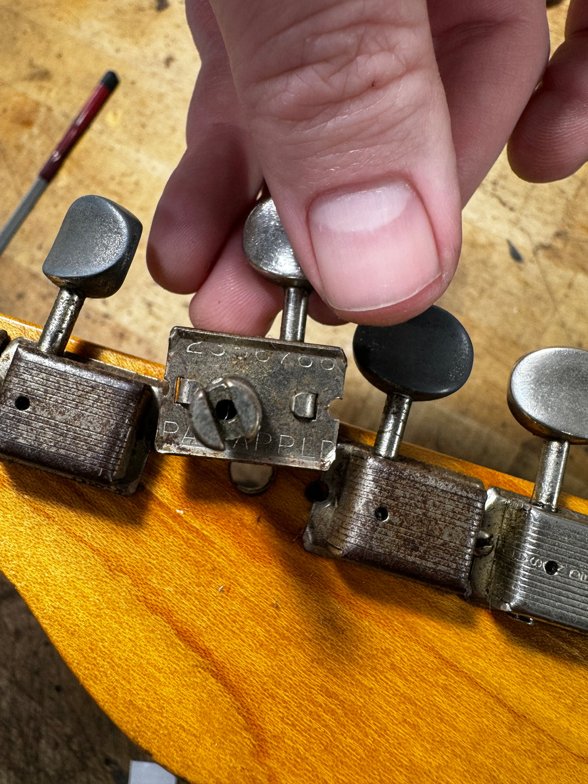 Close-up of a hand adjusting a worn, tarnished metal tuning machine on a butterscotch blonde headstock, showing a black tuner button.