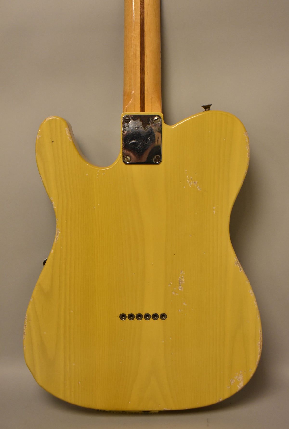 The back of a butterscotch blonde electric guitar, showing a maple neck, chrome neck plate, six string ferrules, and heavy finish wear.