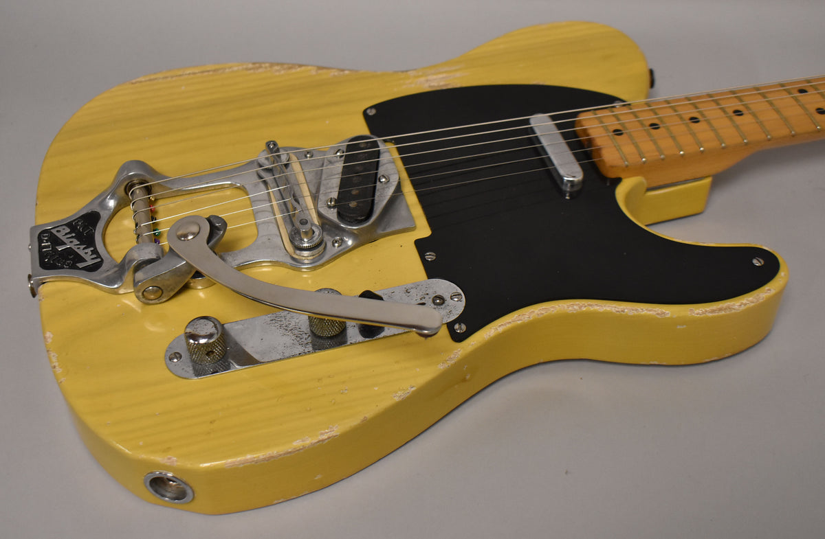 Angled view of a butterscotch blonde guitar with heavy wear, a black pickguard, a Bigsby vibrato, and two knurled chrome knobs.