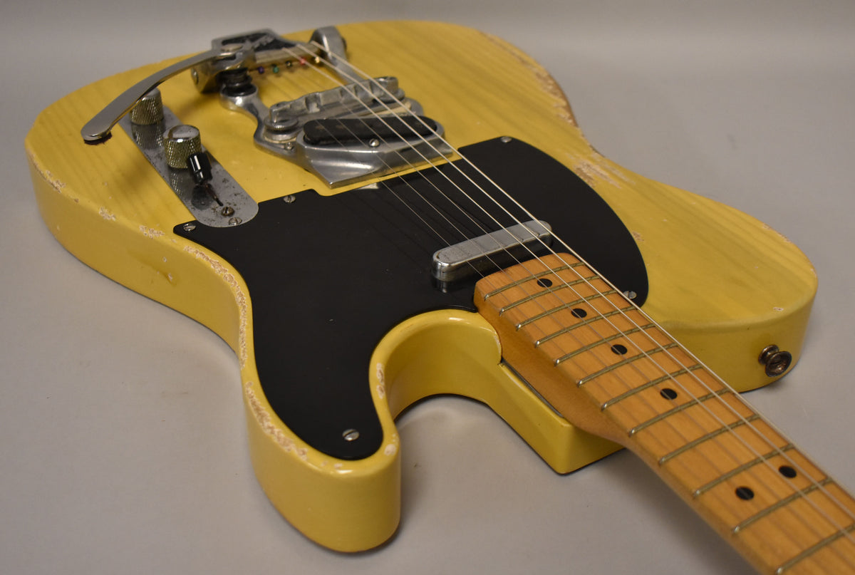 Close-up of a distressed butterscotch blonde electric guitar with a black pickguard, Bigsby tremolo, and maple neck.
