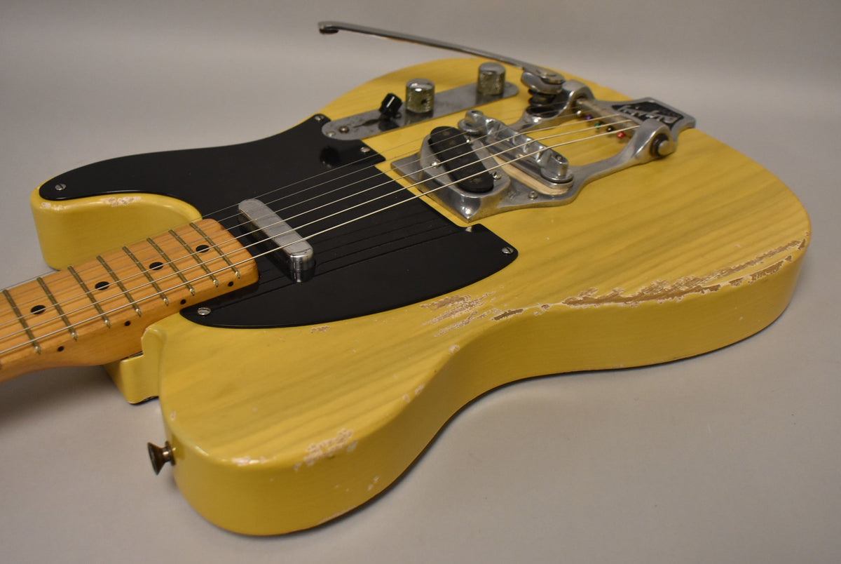 The butterscotch blonde guitar body, with a black pickguard and Bigsby vibrato, shows significant wear and paint chipping along its edges.