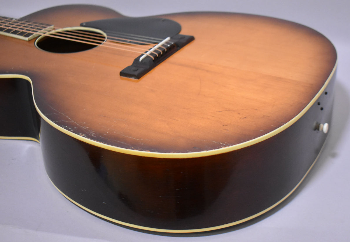 Close-up of the sunburst acoustic guitar's lower bout, showing a black pickguard, bridge, and strap button, with visible finish wear.