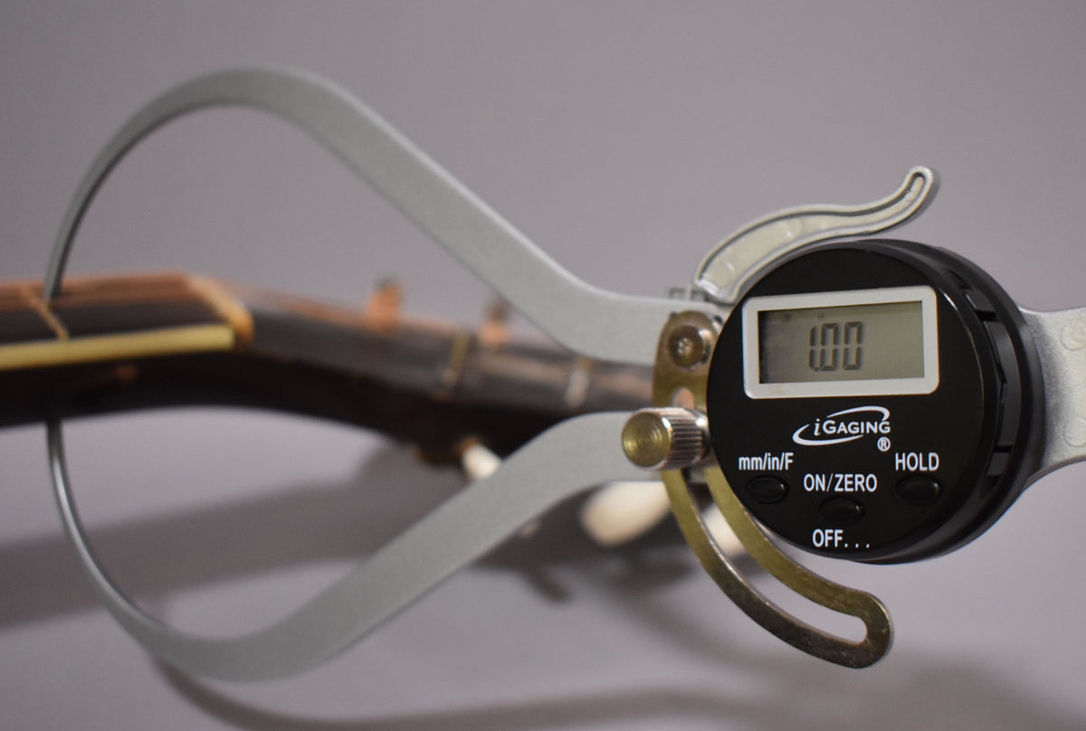 A digital caliper measures the dark brown fretboard, showing "1.00" on its screen, with a light-colored nut visible.
