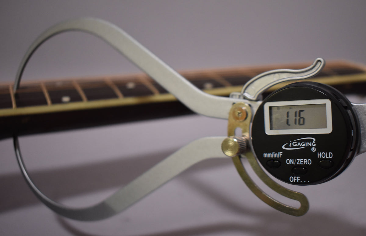 A digital caliper measures the guitar's fretboard, showing "1.16" on its screen, with the dark wood and light binding visible.