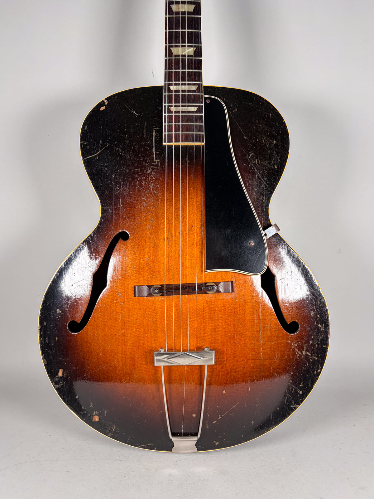 Full front view of a sunburst archtop guitar with heavy finish checking, a black pickguard, and trapezoid fretboard inlays.