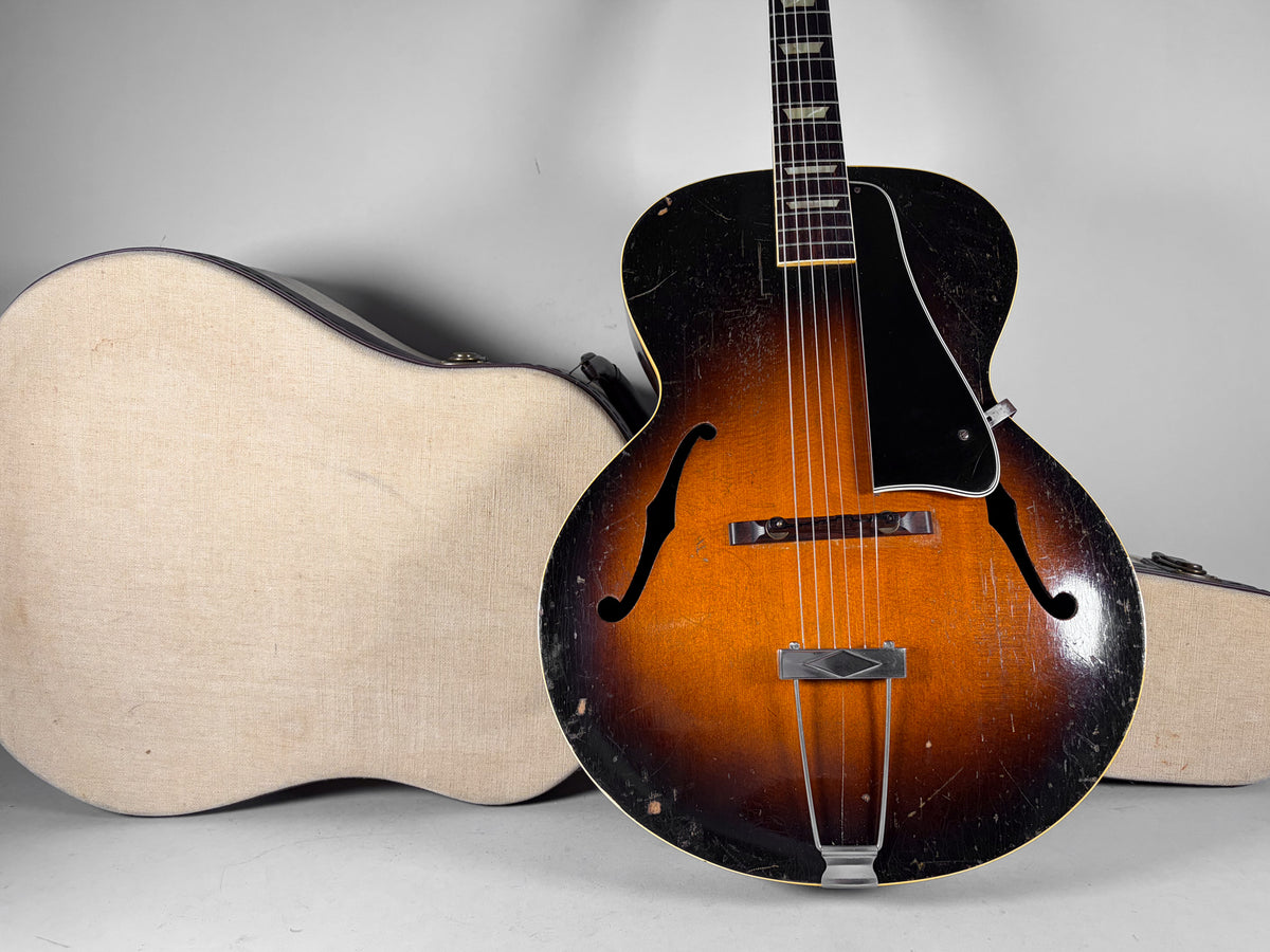 Sunburst archtop guitar with heavy finish wear, black pickguard, and trapezoid tailpiece, next to a light-colored hard shell case.