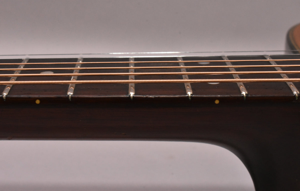 Close-up of the dark fretboard and frets, showing light wear and a small chip between the 5th and 6th frets, with bronze strings.