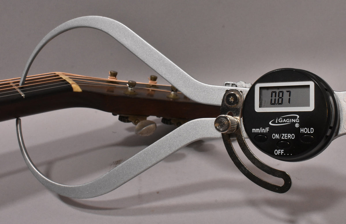 A digital caliper measures the guitar's neck thickness at 0.87 inches, showing the dark brown neck and headstock with tuning pegs.