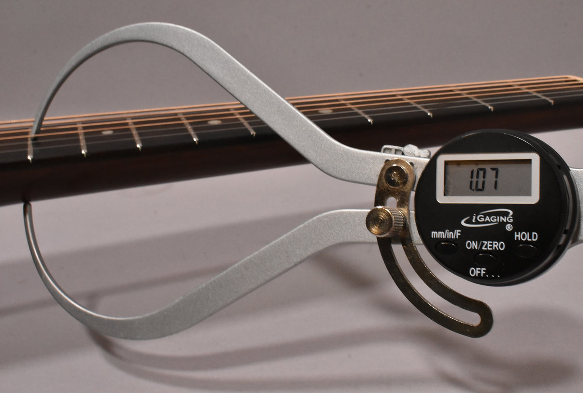 A digital caliper measures the guitar's dark fretboard and strings, displaying "1.07" on its screen.