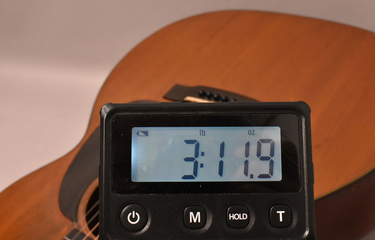 A digital scale displaying "3:11.9 lb oz" in the foreground, with the natural finish acoustic guitar body blurred behind it.