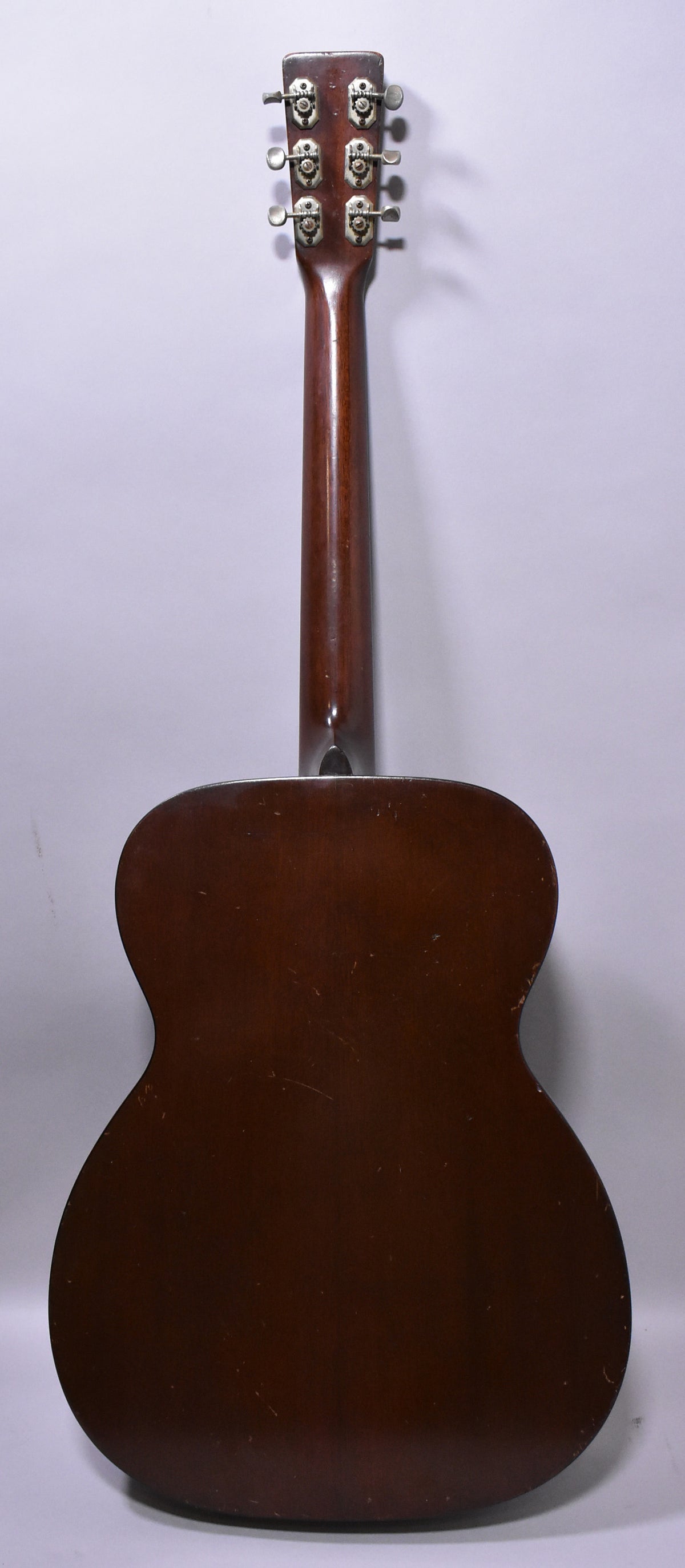 The back of a dark brown acoustic guitar, showing the body, neck, and headstock with six metal tuners, all with visible wear and scratches.