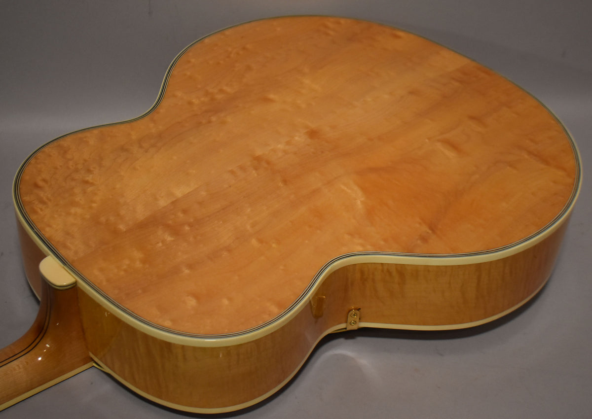 The natural archtop acoustic guitar's back and sides show a light wood grain with subtle figuring and cream binding.